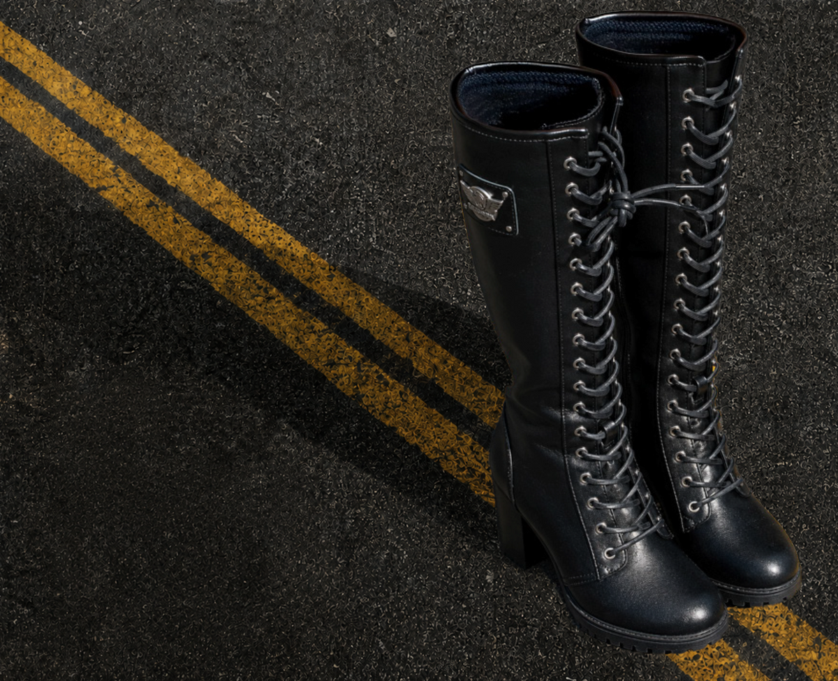 A pair of black knee-high lace-up boots with a slight heel, photographed standing on dark asphalt with double yellow road lines visible.